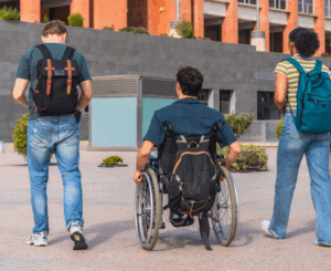 A imagem apresenta três adolescentes andando lado a lado. À esquerda um rapaz usando calça jeans, blusa verde e uma mochila, ao centro um rapaz na cadeira de rodas e com uma mochila e à direita uma adolescente usando jeans e blusa com listras brancas e amarelas e uma mochila verde. Ao fundo um prédio de uma escola.